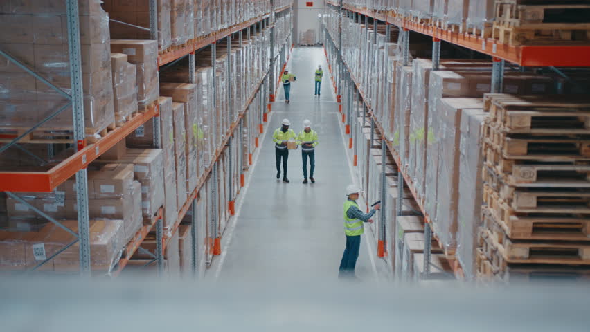 Hardworking warehouse employees wearing safety helmets and vests walking between high pallet racks. Men scanning items with devices. Workers transporting boxes or checking inventory data on tablet.