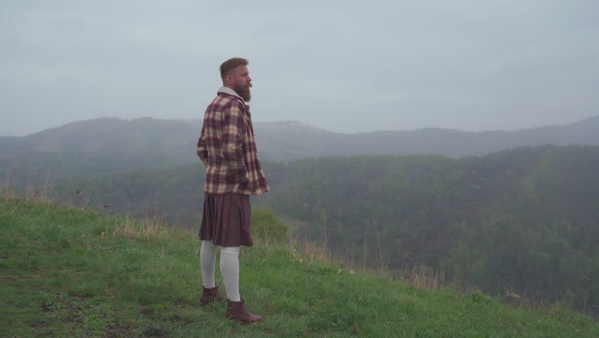 An adult redhead bearded man in Scottish national dress plaid jacket and kilt stands on a hill and looks into the distance. Masculinity, pride and human dignity concept.