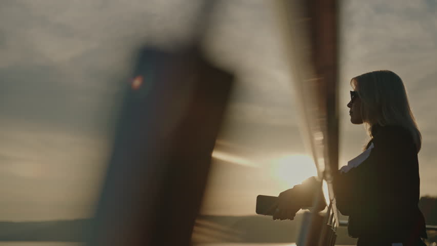 Silhouette of stylish woman adjusting sunglasses, holding smartphone, leaning on railing of pier, overlooking serene lake and rolling hills during golden sunset landscape. Shooting in slow motion.