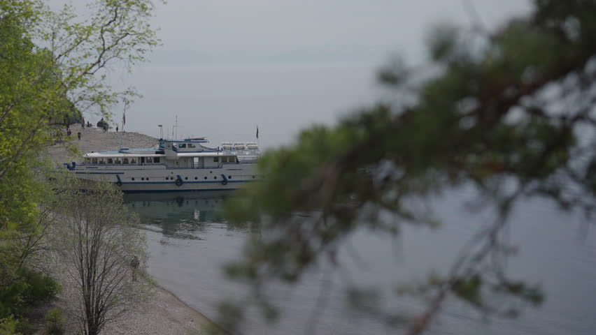 View from mountain to passenger ferry docking at shore of lake Baikal with tourists waiting on pier, framed by lush greenery in serene natural setting. Concept of vacation and travel in Russia.