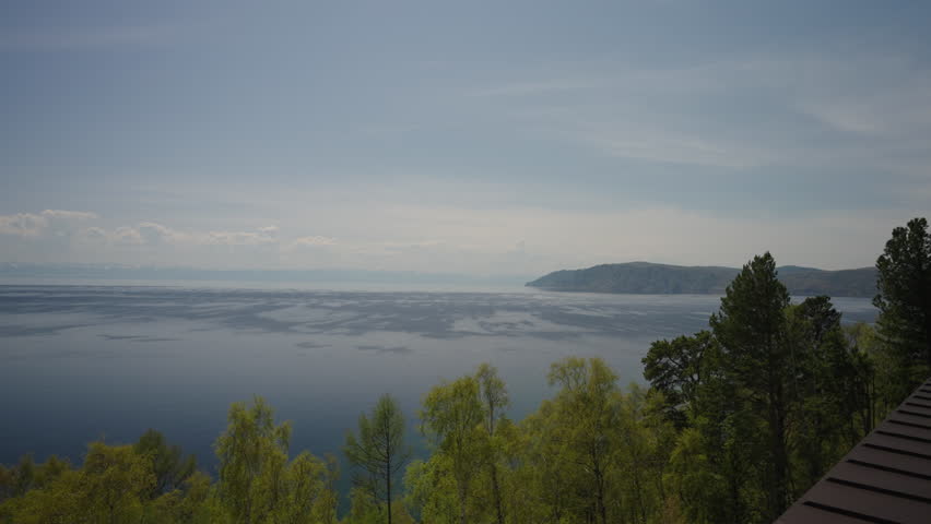 Tranquil scene of lone boat sailing on vast expanse of lake Baikal at dusk, with shoreline trees framing view and cloudy sky overhead creating serene atmosphere, no people, nobody, slow motion.