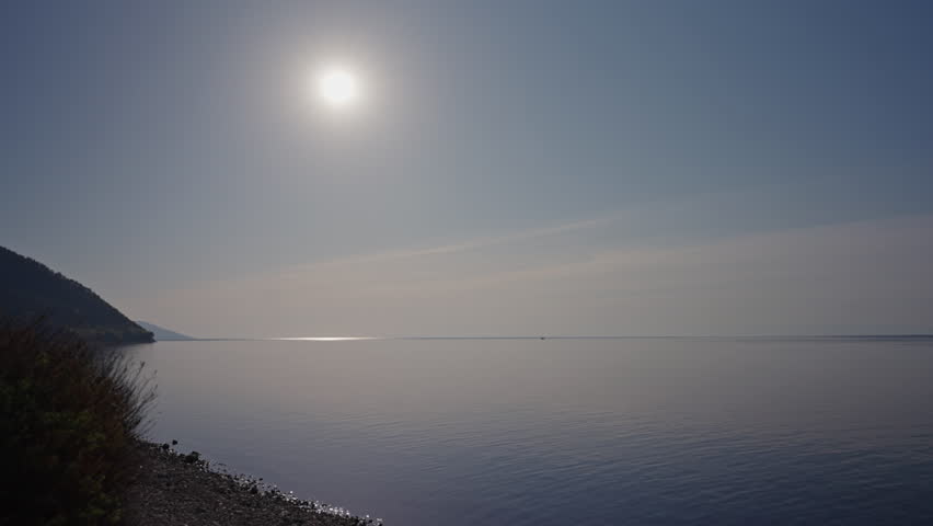 Serene view of sunlight over rocky lake Baikal shoreline, illuminating verdant vegetation and towering mountain peaks against blue sky, nobody, slow motion. Concept of vacation and travel in Russia.