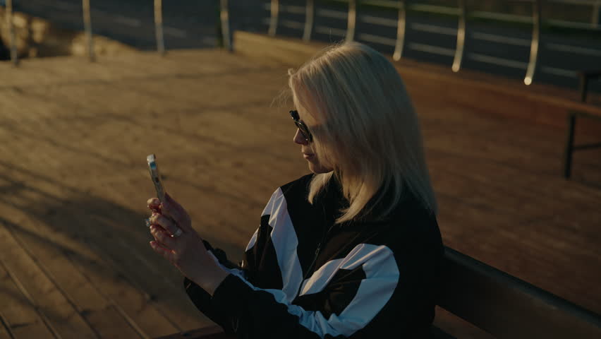Stylish young blonde woman taking pictures or recording videos using phone during beautiful golden hour sunset while sitting on bench in pier. Concept of vacations and travel. Shooting in slow motion.