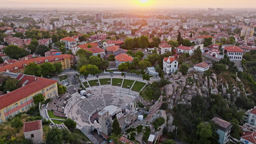 Plovdiv sunrise drone view highlighting Roman Theatre nestled among hills, red roof houses, historic city Bulgaria, amphitheater cultural landmark attracting travelers, warm summer light capturing 