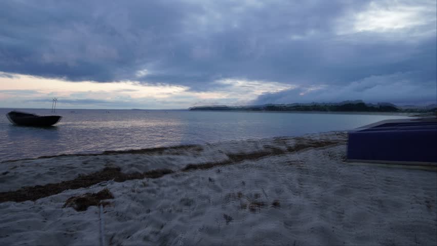 A time-lapse scene of dusk over a quiet beach shows drifting boats, dark clouds, and fading coastal light