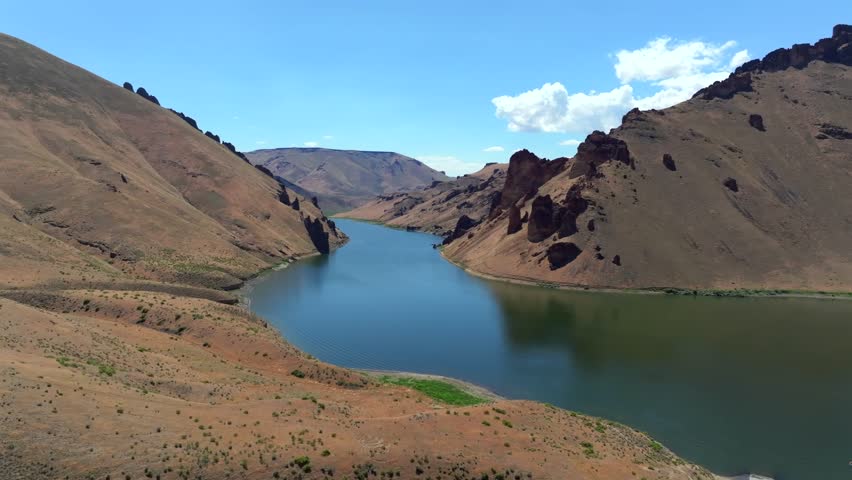 An aerial shot presents Leslie Gulch Lake cutting through a vast, arid canyon, flanked by steep, brown mountains under a blue sky in Oregon
