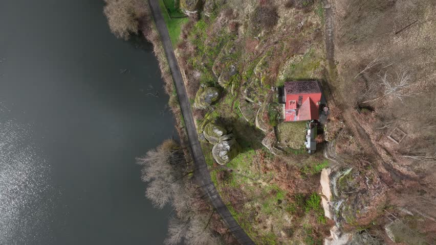 An aerial view of a red-roofed cottage on the rocky shore of Rybnik Vezak in Krckovice, Czech Republic, surrounded by early spring forest