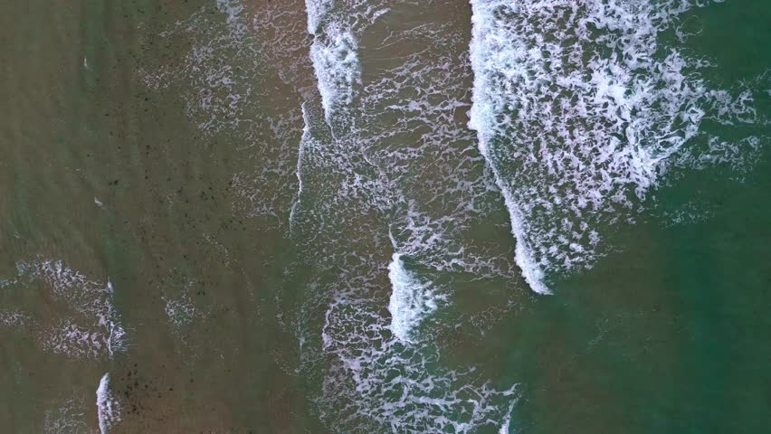 Aerial beach waves crashing gently on sandy shore with rhythmic motion.
