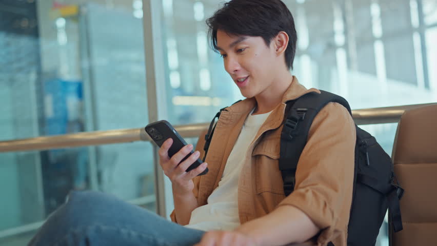 Happy young Asian man waiting for departure at the airport terminal while using his phone, social media, internet