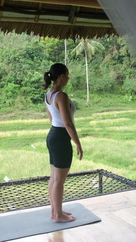 Vertical video. Woman doing yoga on bamboo terrace surrounded by breathtaking tropical rice fields, slow motion 