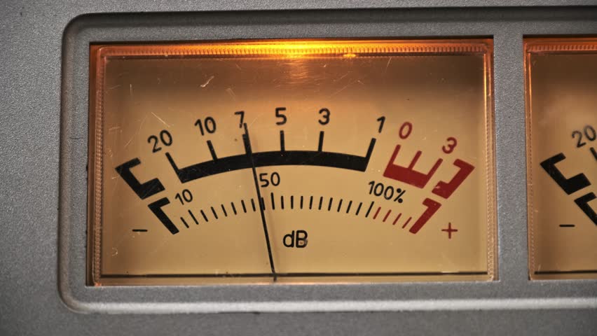 Static macro shot of a brightly lit analog VU meter. The decibel scale needle actively moves, measuring audio output on a vintage tape deck or amplifier, showing peaks and troughs.