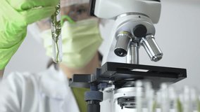 Scientist wearing green gloves and a face mask selecting a test tube with transparent liquid from a rack near a microscope in a lab. Medicine, healthcare and science concept - Powered by Shutterstock - Get 15% off with code: PIKWIZARD15