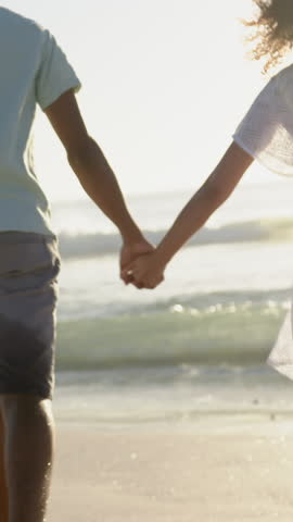 Walking couple wading in shallow surf by shoreline, with interlocked hands and wet sand. Romantic, companionship, seaside, vibrance, casual, adventure, harmony