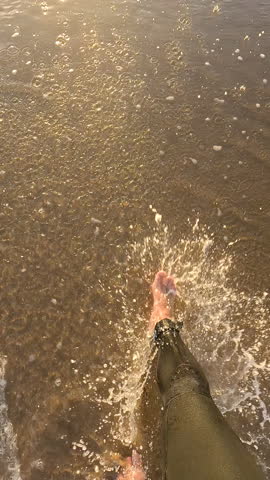 VERTICAL, POV, SLOW MOTION, LENS FLARE: Surfer walks along sandy shoreline and looks up to sunset. Golden light illuminates beach while gentle waves wash ashore and a few people enjoy in the water.