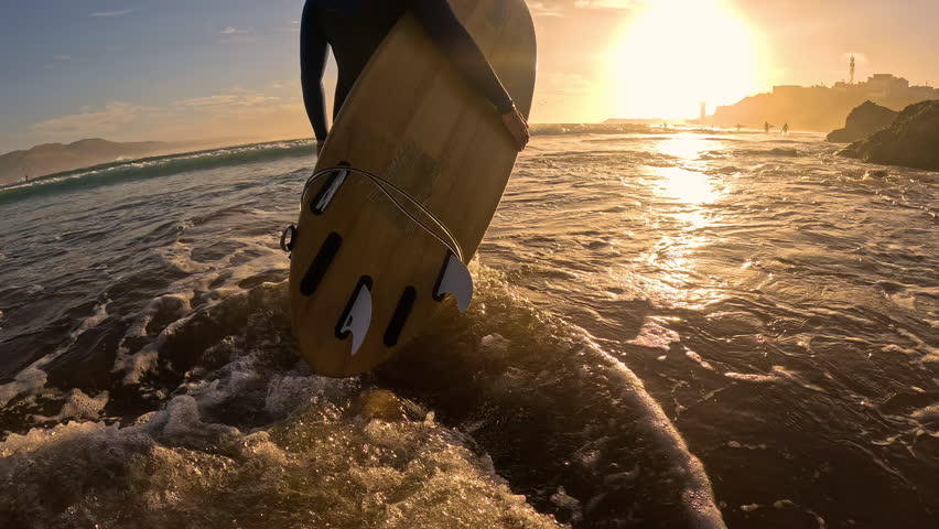 LENS FLARE, SLOW MOTION: Following female surfer with wooden surfboard through shallows at sunset. She came out of water after surf session and is walking along the shore, bathed in golden setting sun