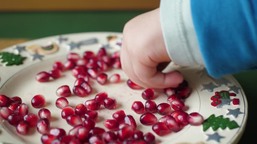 A childs small hand eagerly reaches for bright red pomegranate seeds on a decorative plate, capturing the joy of festive treats enjoyed during family gatherings and celebrations
