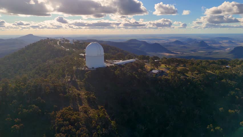 Aerial footage glides over a mountaintop observatory surrounded by forest, with dramatic clouds and warm sunset light illuminating the landscape and distant hills