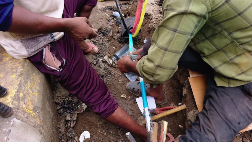 Two laborers mend underground telecoms cables by roadside, Technicians fix communication lines under the curbside, Wire man reestablishes citywide phone circuits, Cords are cut and spliced cautiously