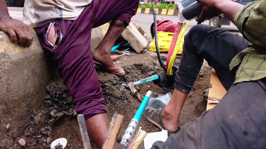 Technicians fix communication lines under the curbside, Two laborers mend underground telecoms cables by roadside, Wire man reestablishes citywide phone circuits, Cords are cut and spliced cautiously