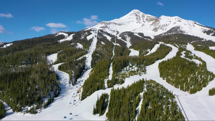 Winter drone view of Big Sky, Montana, features chair lifts packed with skiers, pine trees blanketed in snow, and thrilling downhill ski trails cutting through scenic mountain slopes.