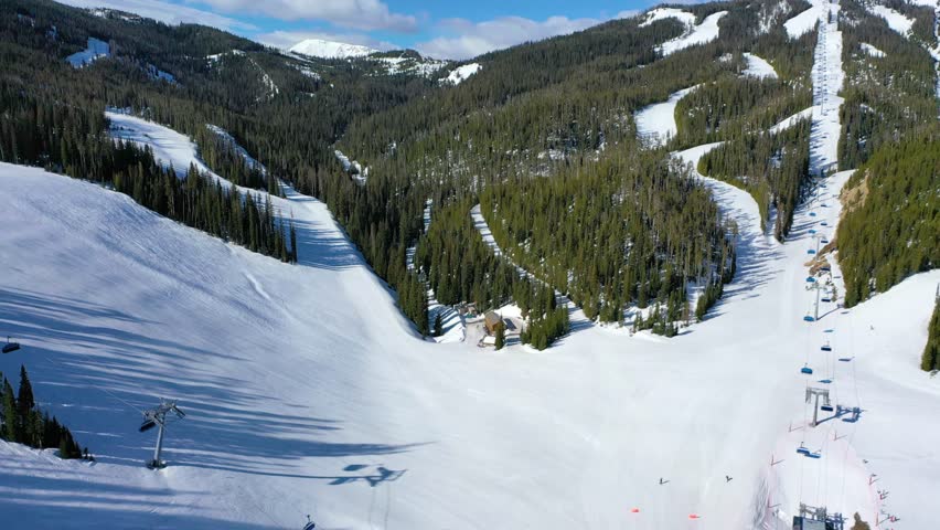 Chair lifts full of bundled skiers cross snowy ridges in Big Sky, Montana, captured by drone over icy runs, pine forests, and powder trails beneath a crisp blue winter sky.