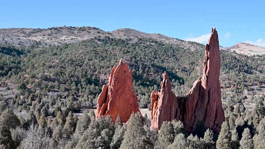 Aerial view of jagged red sandstone hoodoos rising from a dense forested landscape with rolling hills in Garden of the Gods, Colorado.