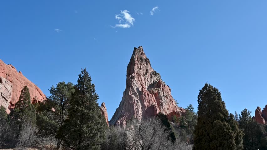 Drone shot of towering red sandstone spire surrounded by pine trees beneath a blue sky at Garden of the Gods in Colorado Springs.