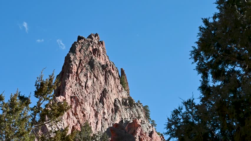 Low drone angle of rugged red sandstone pinnacle framed by treetops and set against a bright blue sky at Garden of the Gods, Colorado.