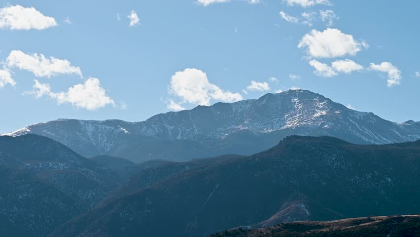 Sweeping drone view of Pikes Peak mountain range under cloudy blue skies, showcasing rugged alpine terrain near Colorado Springs.