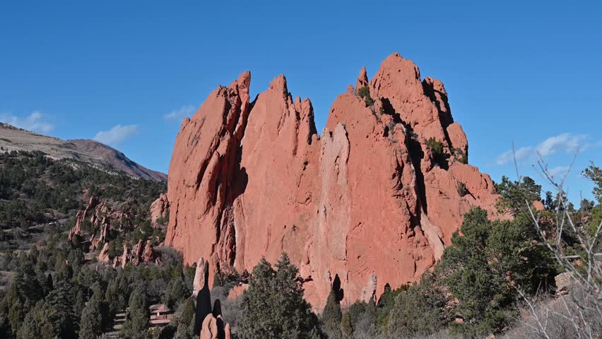 Drone shot of sunlit red sandstone monoliths surrounded by pine trees and distant hills at Garden of the Gods in Colorado Springs.