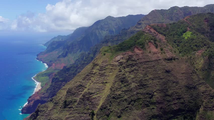 Smooth, parallax, 4k drone shot of Na Pali Coast ridges from the Awa'awapuhi trail in Waimea Canyon State Park in Kauai. See the white puffy clouds, lush green ridges, and turquoise water below.