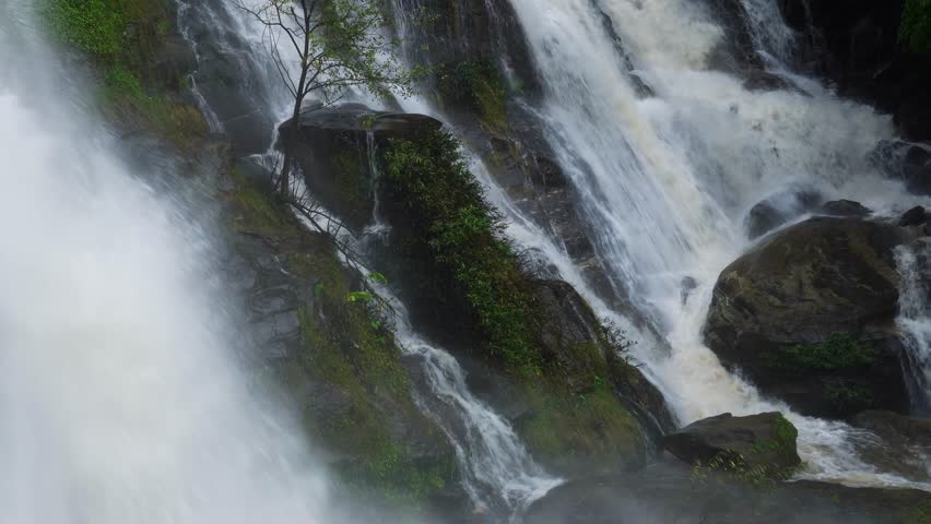 A strong waterfall cascading through a lush green forest during the rainy season. The river current is powerful and the water has turned muddy after heavy rainfall, showcasing the raw energy of nature