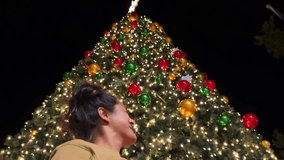 Young woman gazing upward at large decorated Christmas tree during nighttime, slow motion capturing magical moment of festive wonder and holiday spirit - Powered by Shutterstock - Get 15% off with code: PIKWIZARD15