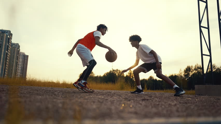Energy of motivated sportspersons, two teens playing basketball, active sport. Young black guys practicing feints in streetball, cinematic slow motion shot, active leisure of modern adolescents