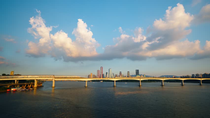 A stunning aerial view shows a metro train crossing the Dangsan Railway Bridge over the Han River in Seoul, with the modern skyscrapers of the Yeouido business district in the background at sunset.