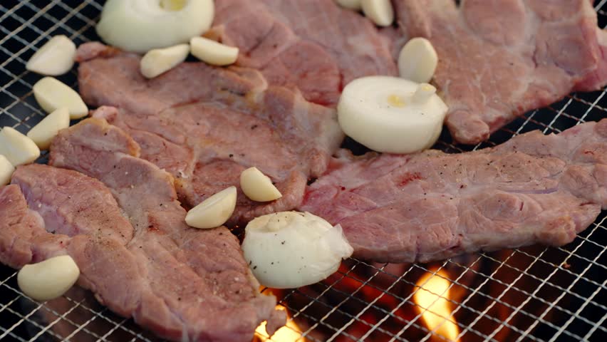 An extreme close-up shot captures seasoned pork slices, garlic cloves, and onions sizzling on a hot barbecue grill over charcoal flames, a popular Korean-style outdoor meal.