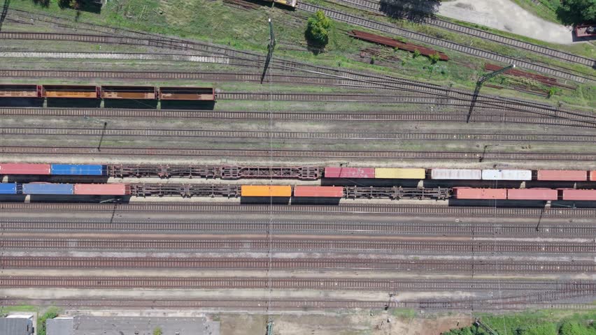 A top-down aerial video showing a freight train loaded with colorful cargo containers in a large railway yard. Multiple train tracks and empty wagons are visible.