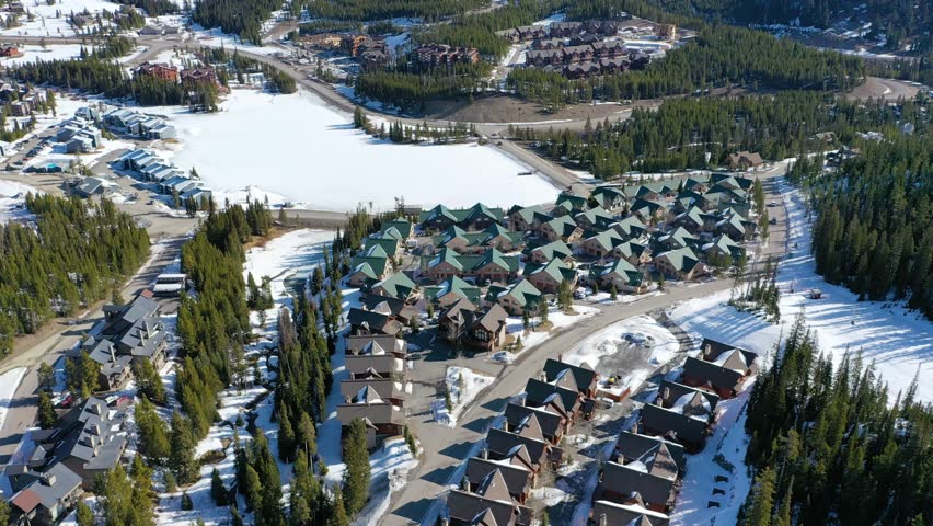 Bird’s eye drone view of Big Sky, Montana shows clusters of vacation condos and snowy streets set among forested hills and framed by dramatic peaks in a peaceful alpine landscape.