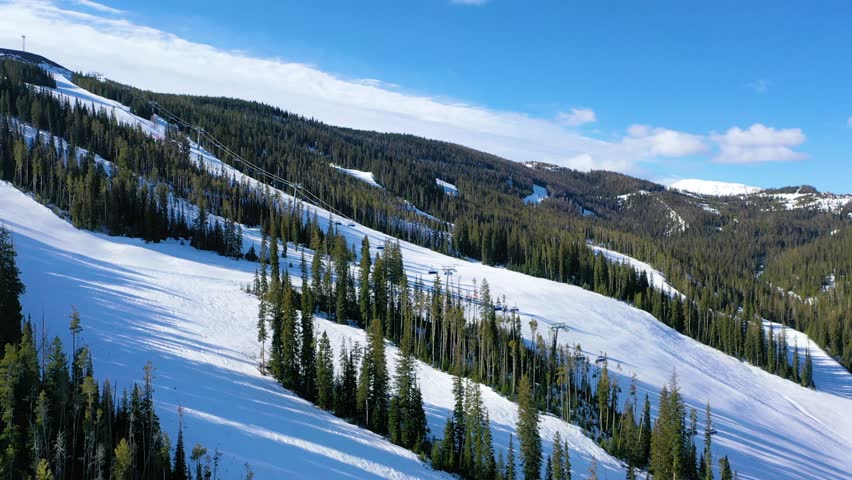 Drone captures a snowy day of skiing at Big Sky, Montana with chair lifts transporting skiers up the slopes, scenic pine forests, and white mountains filling the winter landscape.