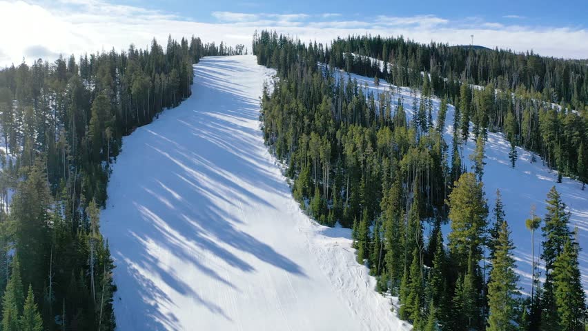 Aerial drone shot flying over a snowy mountain in winter, showcasing scenic ski trails winding through pine trees, with panoramic alpine views and a crisp, refreshing mountain atmosphere.