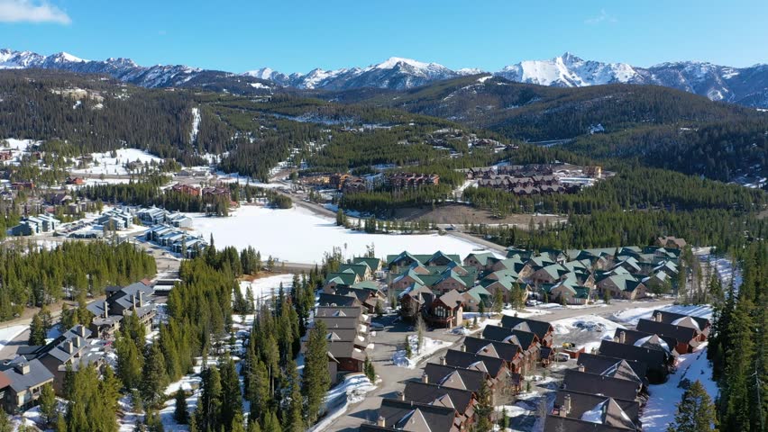 Overhead drone view captures the town of Big Sky, Montana blanketed in snow, with charming vacation condos, frosted roads, and pine-covered hills beneath towering mountain peaks.
