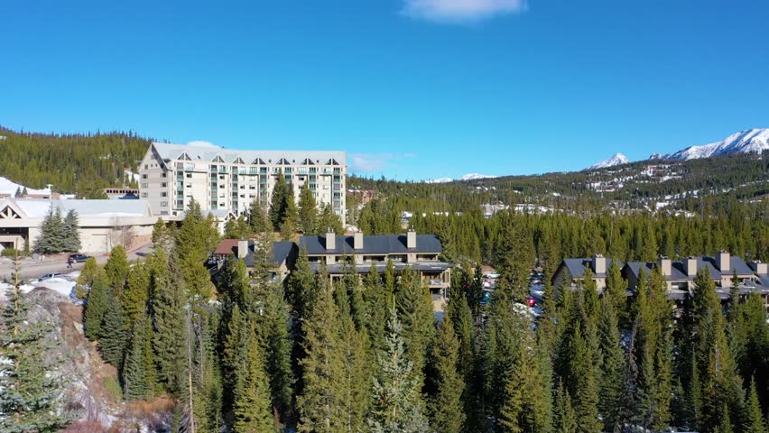 Aerial drone footage captures a packed parking lot in snowy Big Sky, Montana, just below the ski resort, with surrounding vacation condos, tall pines, and vast mountain scenery all around.