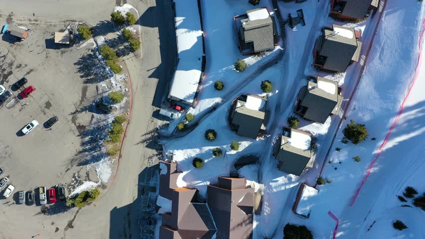 High drone shot of Big Sky Montana condos in a snow-covered mountain town, with frozen roads, smoke rising from chimneys, and peaceful woods surrounding this idyllic alpine retreat.