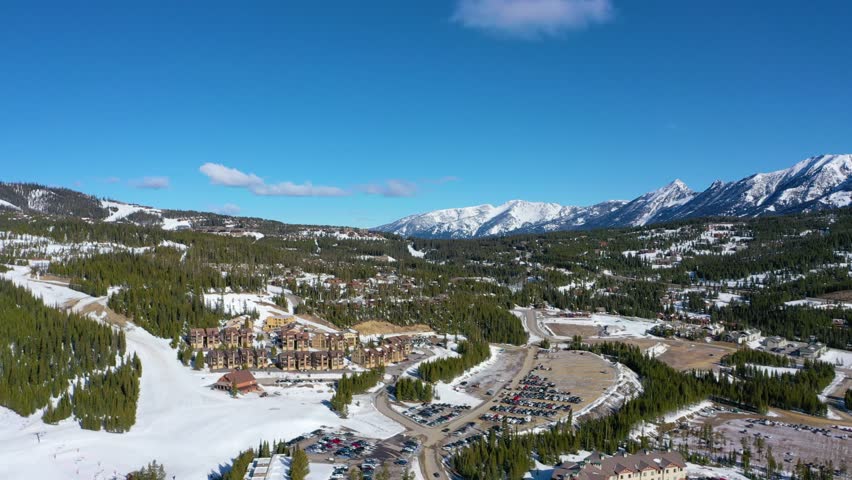Drone soars above a busy winter parking lot near the heart of Big Sky, with snow-draped condos, icy roads, and distant peaks just steps from the base of the mountain resort.