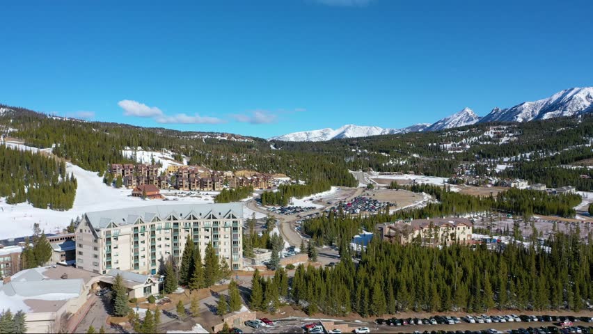 Aerial view reveals cars tightly packed into a snowy lot in Big Sky, Montana, framed by pine trees, rental condos, and towering alpine peaks just minutes from the nearby ski resort.