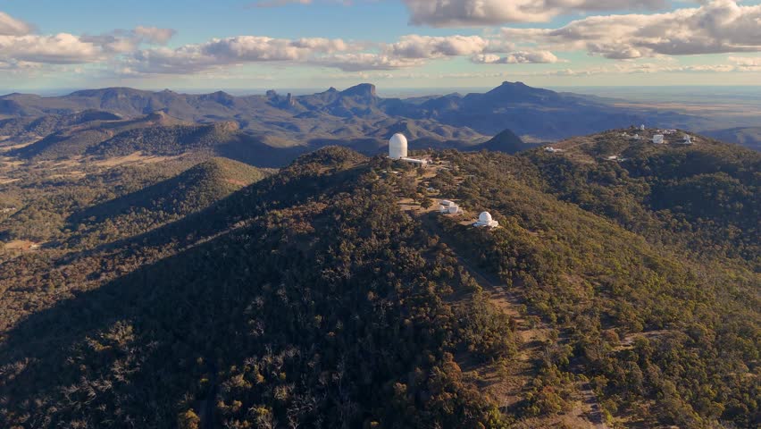Aerial drone footage glides over a forested mountain ridge, revealing a large observatory dome and telescopes under warm, late afternoon sunlight