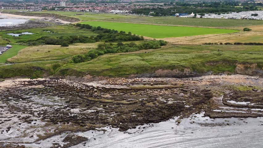 Drone camera glides above a rocky shoreline and lush green fields, revealing coastal grassland, farmland, and distant town under soft daylight