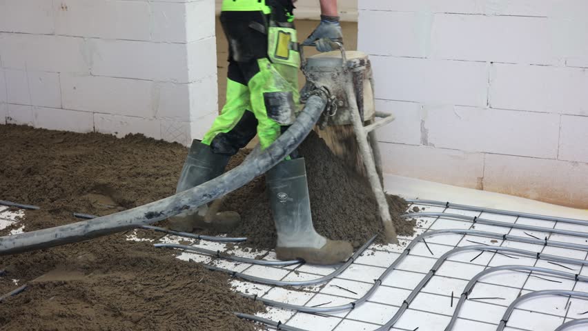 Worker Pouring Concrete Floor in New Building, Construction worker in safety gear pouring concrete over steel reinforcement mesh inside unfinished building, preparing for floor installation.