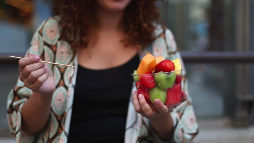 Young woman savoring colorful fruit salad while sitting outdoors, enjoying fresh, vibrant nutrition during summer city lifestyle moment