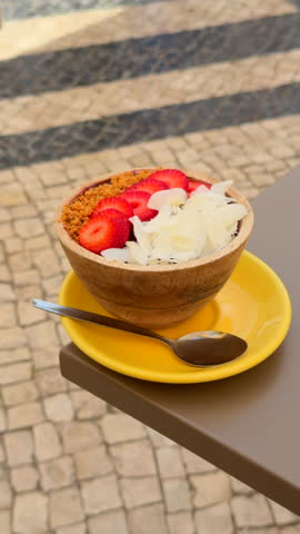 Tropical açai bowl topped with fresh strawberries, coconut flakes and paçoca, served in a wooden bowl on a yellow plate outdoors.